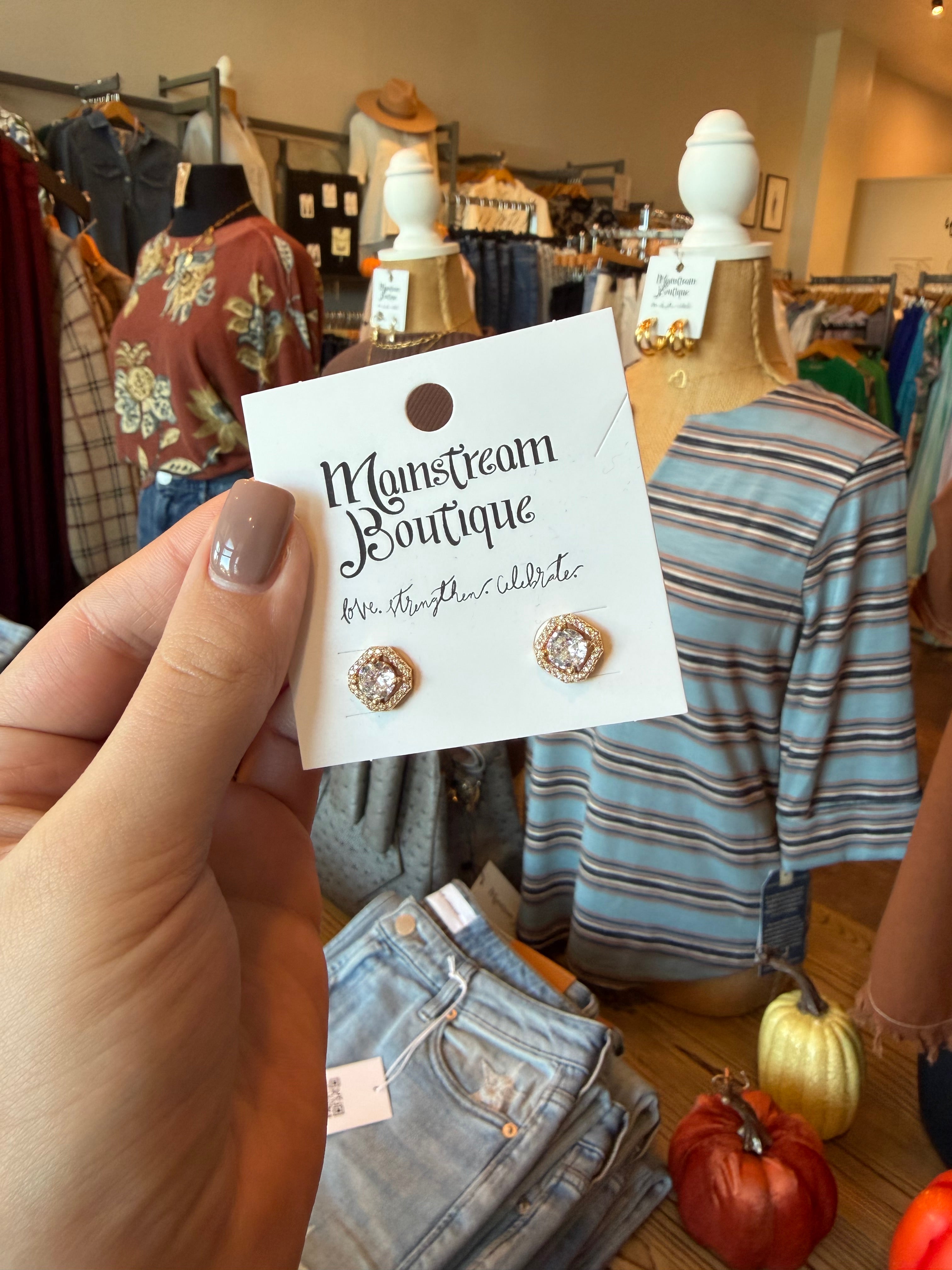 A hand holds a white display card featuring a pair of gold-toned stud earrings with clear stones. The card reads 'Mainstream Boutique' and 'Mr.