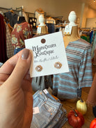 A hand holds a white display card featuring a pair of gold-toned stud earrings with clear stones. The card reads 'Mainstream Boutique' and 'Mr.