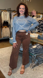 A woman stands in a clothing boutique wearing a light blue cropped sweater with a white heart outline on the chest and brown wide-leg pants. The boutique has clothing racks, mannequins, a wooden table, and hanging chandeliers in the background.