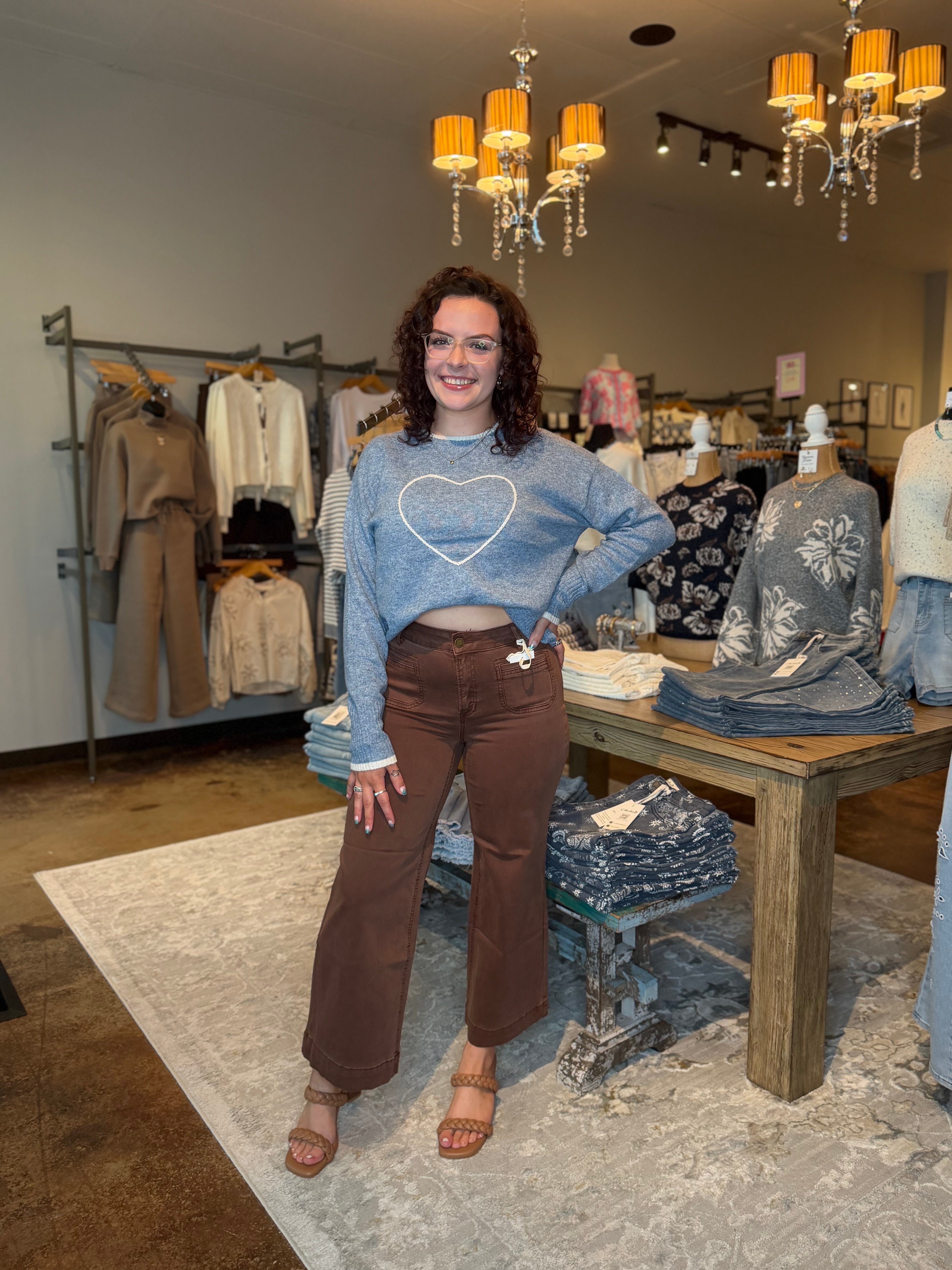 A woman stands in a clothing boutique wearing a light blue cropped sweater with a white heart outline on the chest and brown wide-leg pants. The boutique has clothing racks, mannequins, a wooden table, and hanging chandeliers in the background.