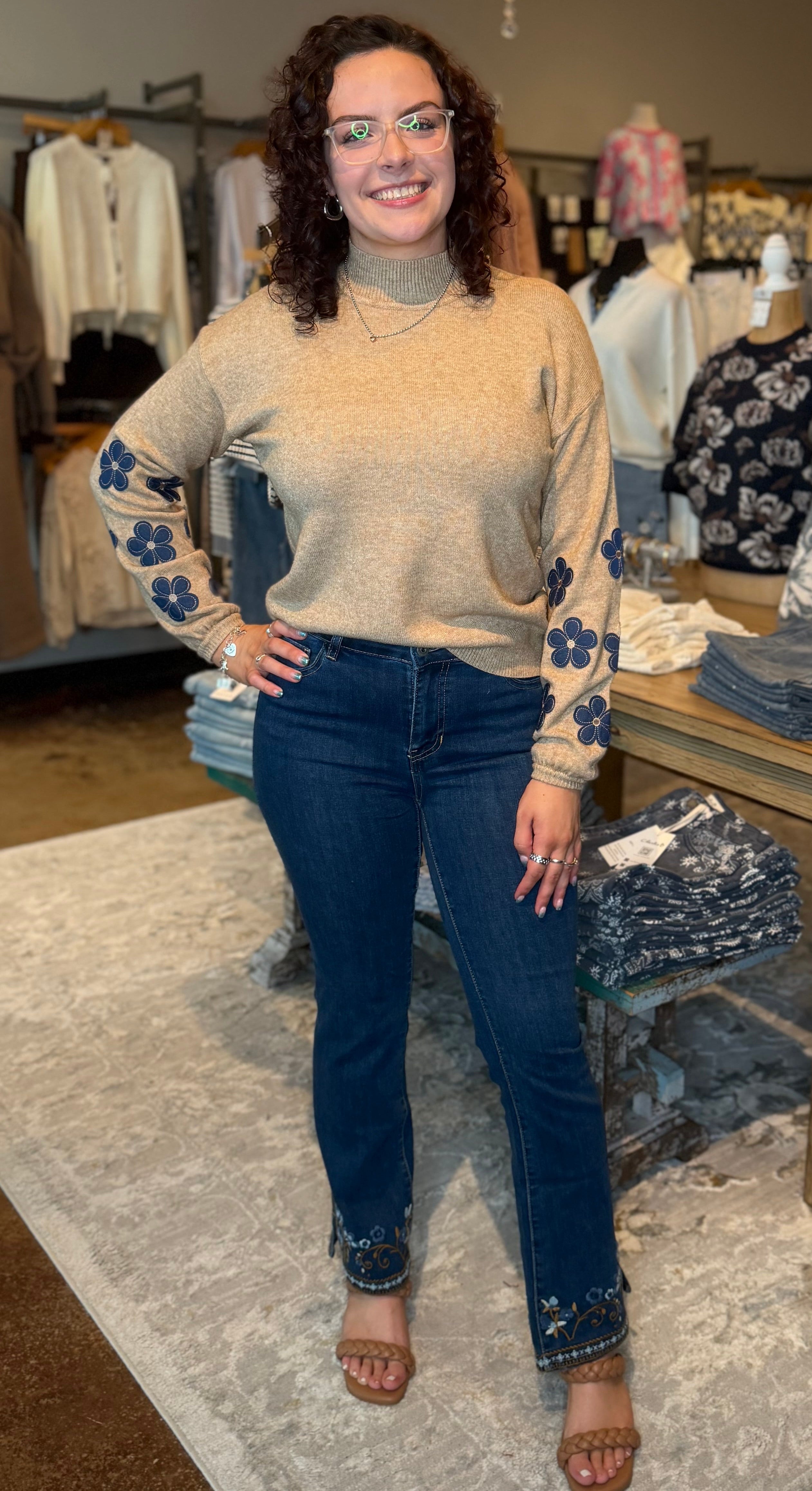 A woman stands in a clothing boutique wearing a beige knit turtleneck sweater with blue floral sleeve details, dark blue jeans with blue floral cuff details, and brown heeled sandals. The boutique has clothing racks, mannequins, a wooden table, and a patterned rug.