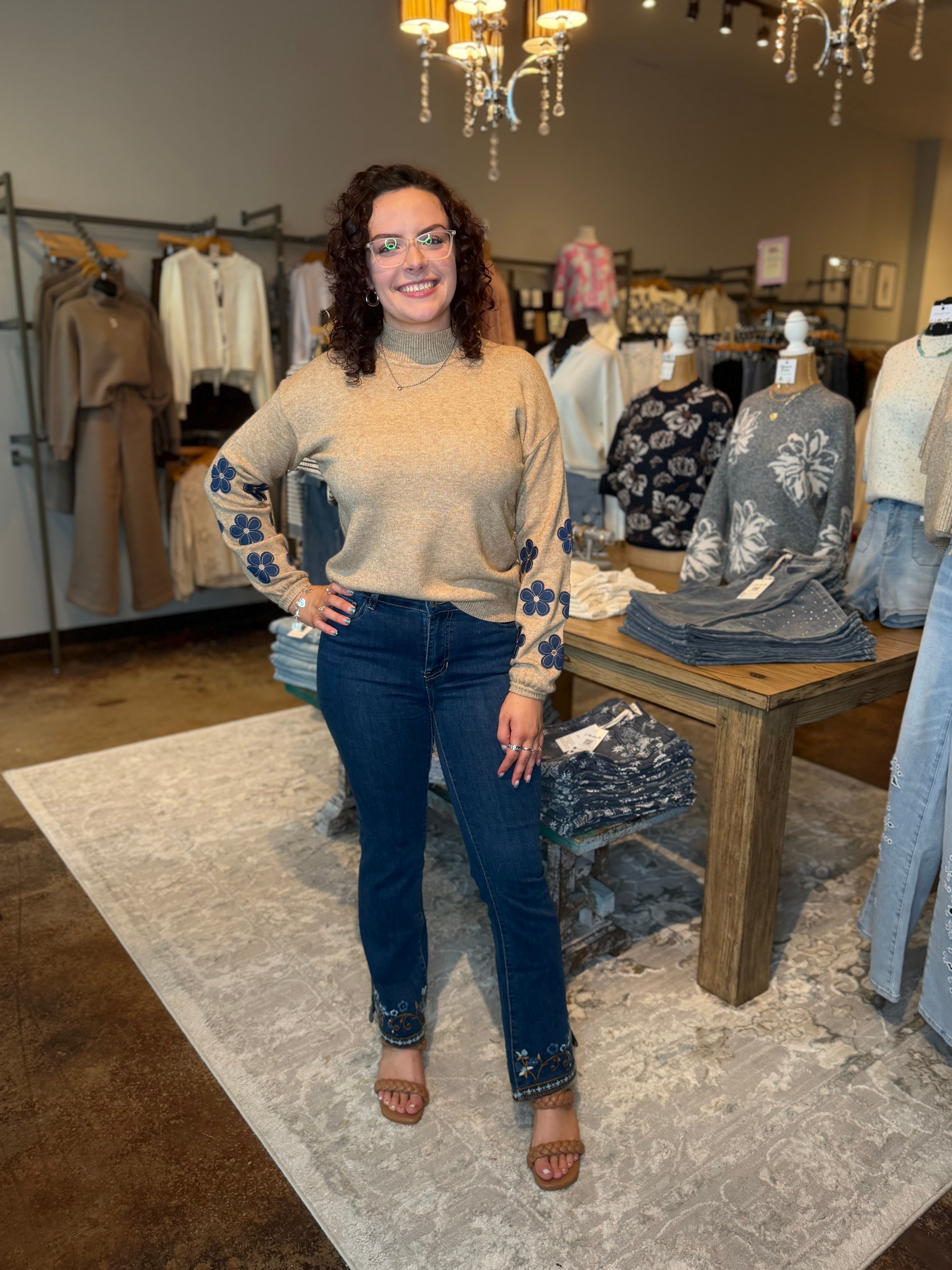 A woman stands in a clothing boutique wearing a beige knit turtleneck sweater with blue floral sleeve details, dark blue jeans with blue floral cuff details, and brown heeled sandals. The boutique has clothing racks, mannequins, a wooden table, and a patterned rug.