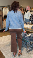 A woman viewed from behind wearing a light blue knit sweater and brown pants, standing inside a clothing boutique near racks of garments and a display table.