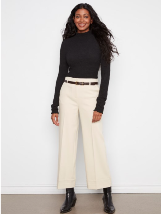 A woman modeling a black long-sleeve mock neck top and cream wide-leg cropped pants with a thin brown belt, paired with black ankle boots, against a white wall.
