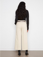 A woman shown from the back wearing a black long-sleeve top, cream wide-leg cropped trousers with a thin brown belt, and black ankle boots, standing against a white wall.
