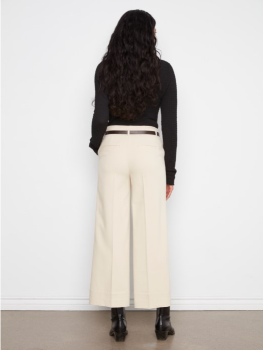 A woman shown from the back wearing a black long-sleeve top, cream wide-leg cropped trousers with a thin brown belt, and black ankle boots, standing against a white wall.