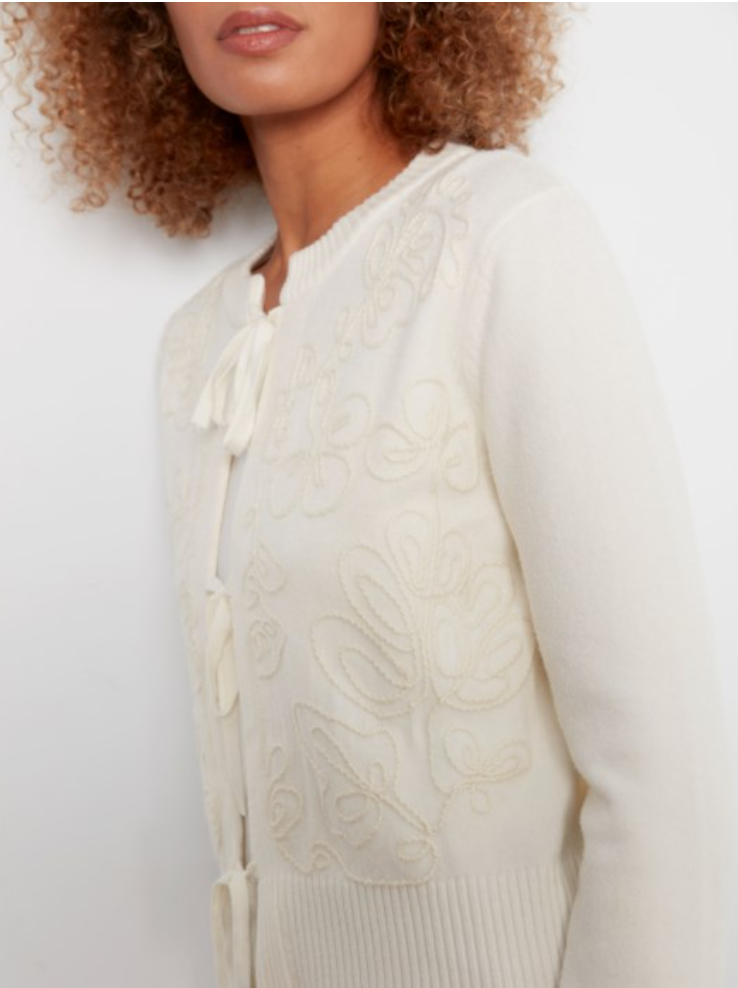 A close-up of a model wearing a cream-colored knit cardigan sweater with matching tie-front buttons and tonal floral embroidery.