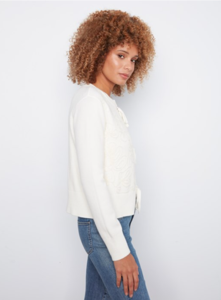 A woman with curly light brown hair is shown in profile wearing a white knit cardigan with lace-up detailing and blue jeans, set against a plain white studio background.