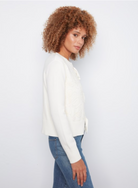 A woman with curly light brown hair is shown in profile wearing a white knit cardigan with lace-up detailing and blue jeans, set against a plain white studio background.
