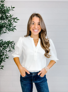 Woman wearing a white blouse and blue jeans standing against a light gray wall with a plant to the left.