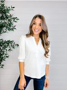 Woman wearing a white blouse and blue jeans standing next to a plant against a light gray wall.
