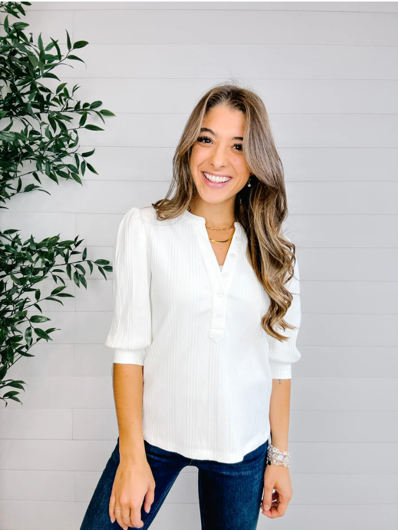 Woman wearing a white blouse and blue jeans standing next to a plant against a light gray wall.