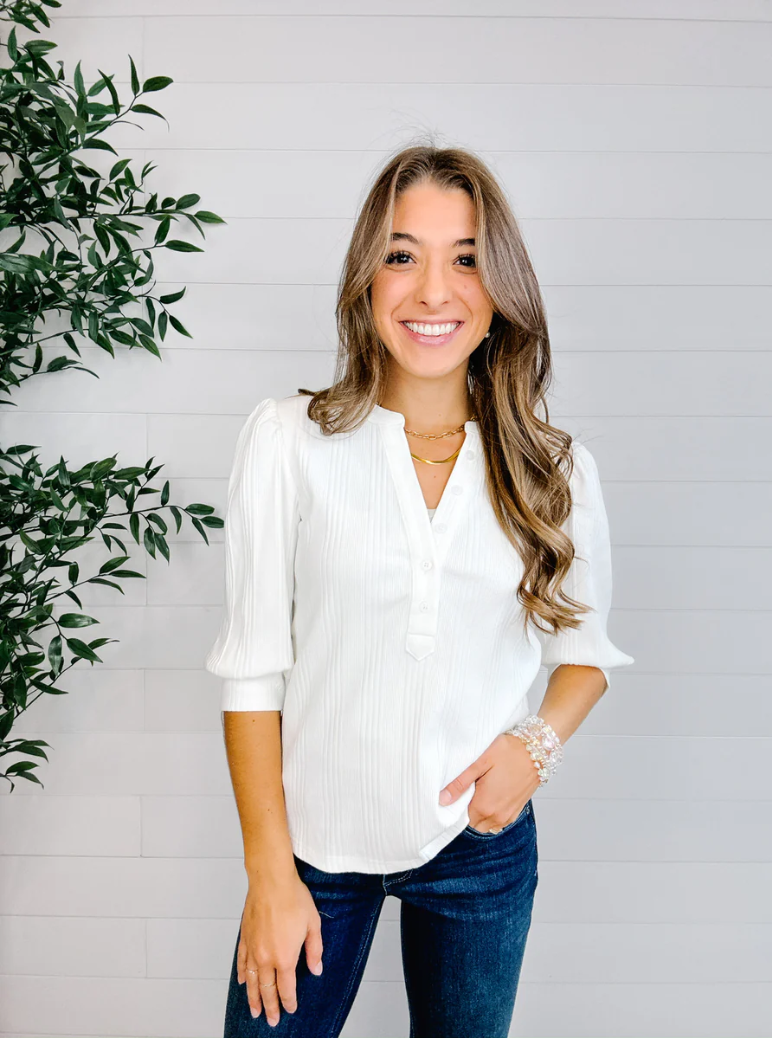 Woman wearing a white blouse and blue jeans standing against a light gray wall with a plant to the left.
