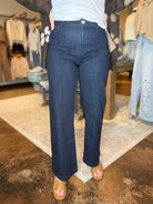 A person wearing dark blue straight-leg denim jeans, a white top, a gray patterned cardigan, and brown sandals, standing in a clothing store.