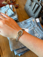 A silver-toned double-band cuff bracelet with a central oval crystal-encrusted setting, worn on a person's wrist. The background shows a wooden table with folded light-wash blue jeans and a gray textured handbag.