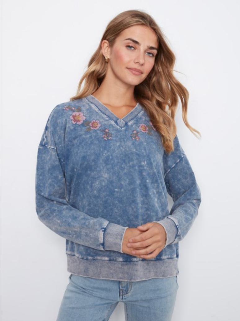 A woman wearing a blue acid-wash V-neck sweatshirt with floral embroidery on the chest, paired with light-wash jeans, against a plain white background.