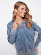 A woman wearing a blue acid-wash long-sleeve sweatshirt with pink floral embroidery on the chest, posed against a plain white background.