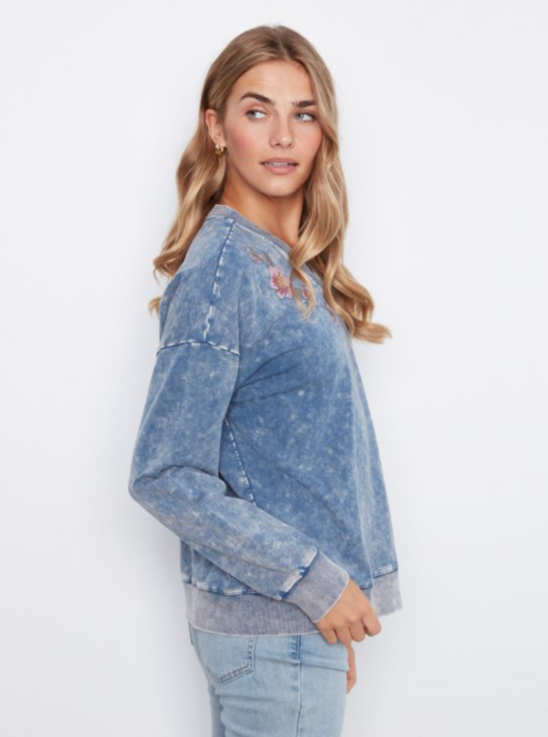 A woman modeling a blue acid-wash long-sleeve sweatshirt with a small embroidered graphic on the chest, paired with light-wash jeans against a plain white background.