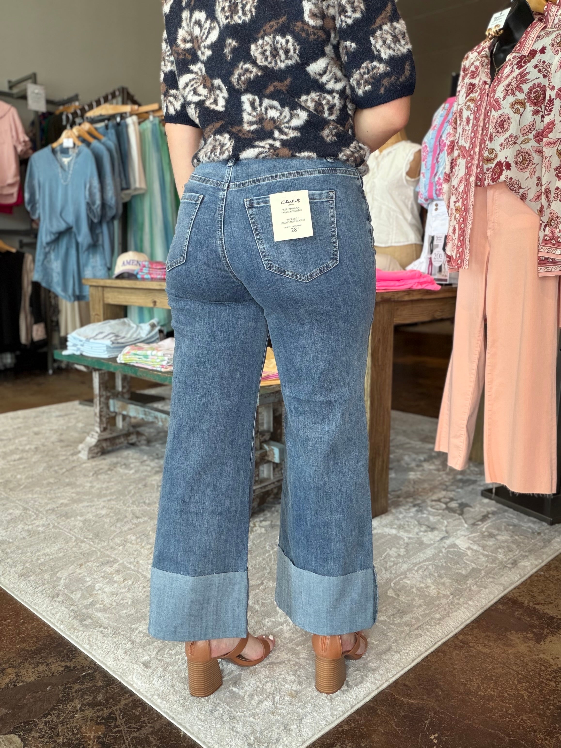 A person shown from the back wearing light-to-medium blue wide-leg denim jeans with rolled cuffs, standing in a clothing store. The jeans have a visible back pocket patch and a white size tag.