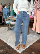 A person standing in a clothing store wearing high-waisted, wide-leg blue jeans with a white long-sleeve top and brown slide sandals. Clothing racks and a patterned rug are visible in the background.