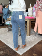 A person standing in a clothing store wearing light-wash blue jeans with a white sweater and brown flat shoes, shown from the back. The jeans have a visible back pocket label.