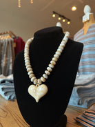A beaded necklace with a heart-shaped pendant displayed on a black mannequin bust inside a clothing store. The necklace features cream-colored beads and a cream-colored heart pendant with a small gold accent.