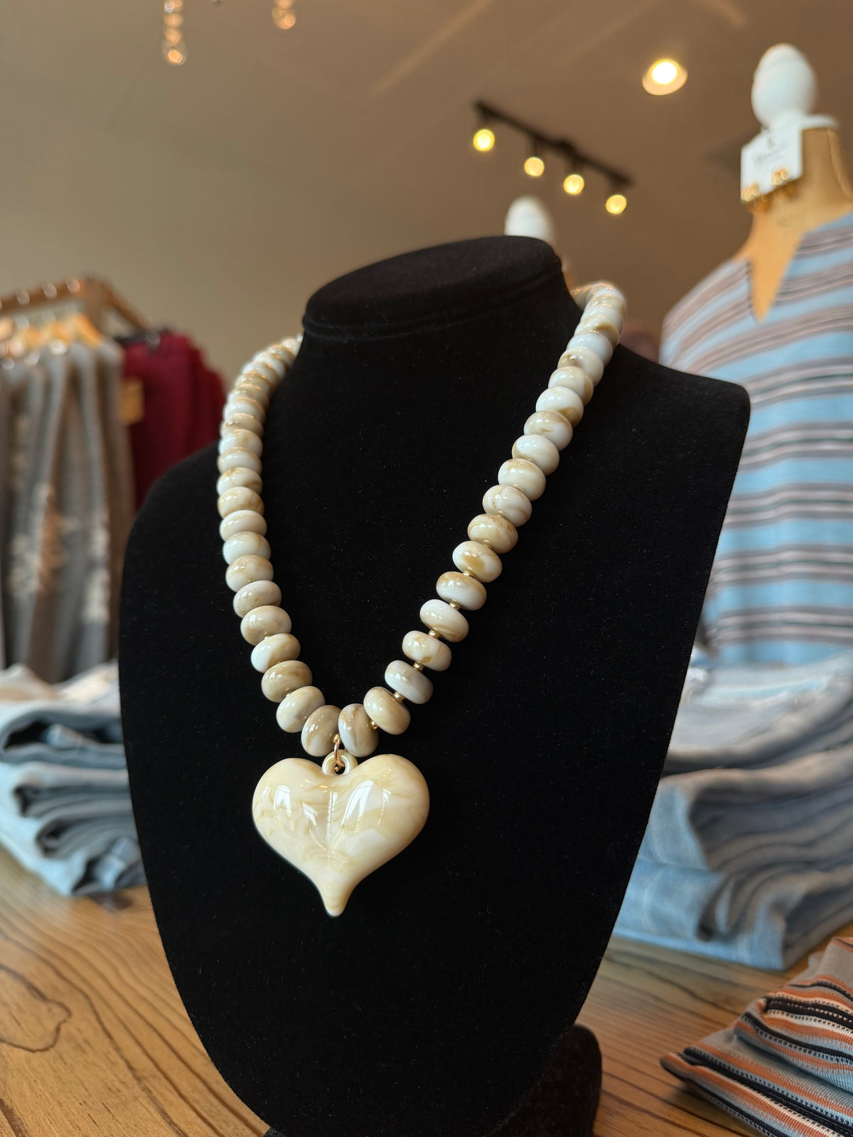 A beaded necklace with a heart-shaped pendant displayed on a black mannequin bust inside a clothing store. The necklace features cream-colored beads and a cream-colored heart pendant with a small gold accent.