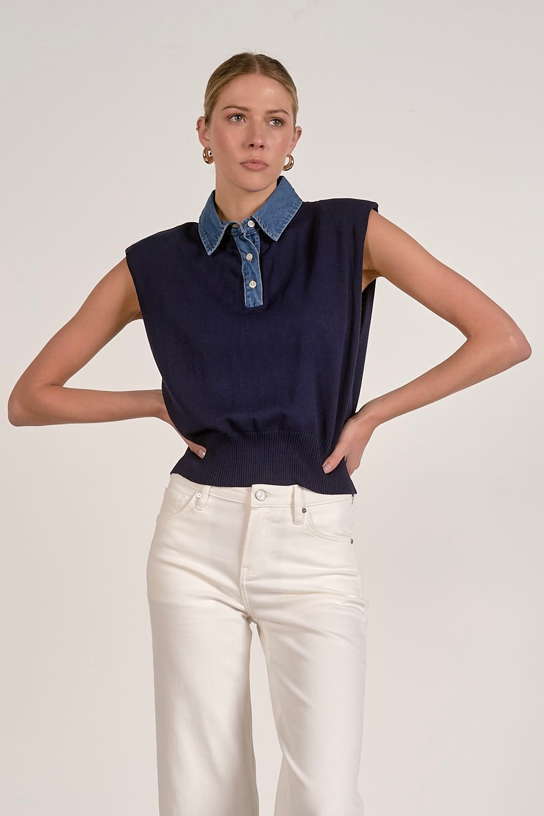 A woman modeling a sleeveless navy knit top with a blue denim collar, paired with white pants, against a plain light gray studio background.