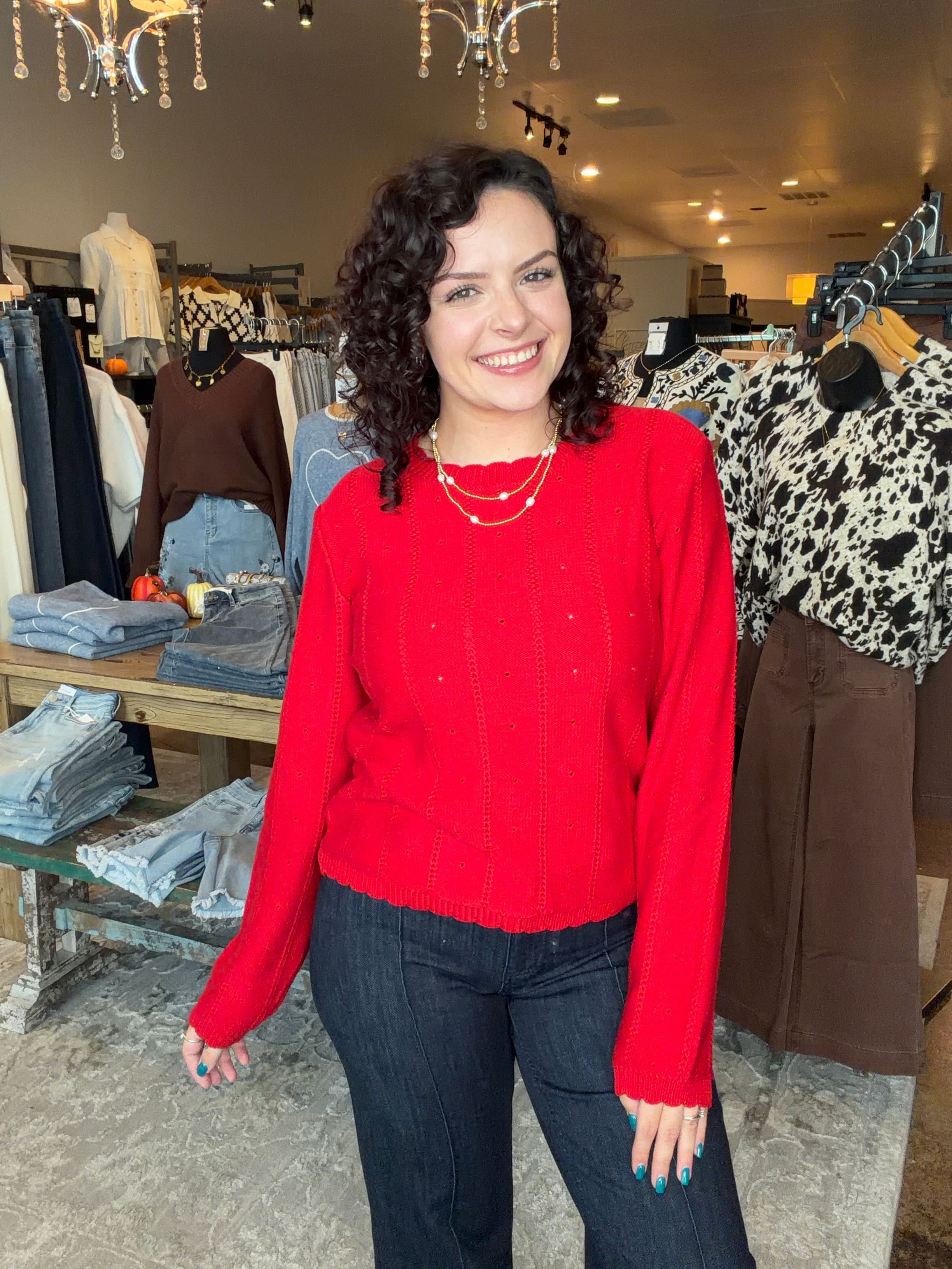 A smiling woman with curly dark hair stands in a clothing boutique wearing a bright red knit sweater and dark jeans. Clothing racks, mannequins, and store fixtures are visible in the background.