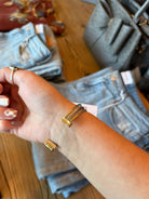 A close-up of a person's wrist wearing a gold-toned wrap bracelet, with folded blue jeans and a gray handbag in the background on a wooden surface.