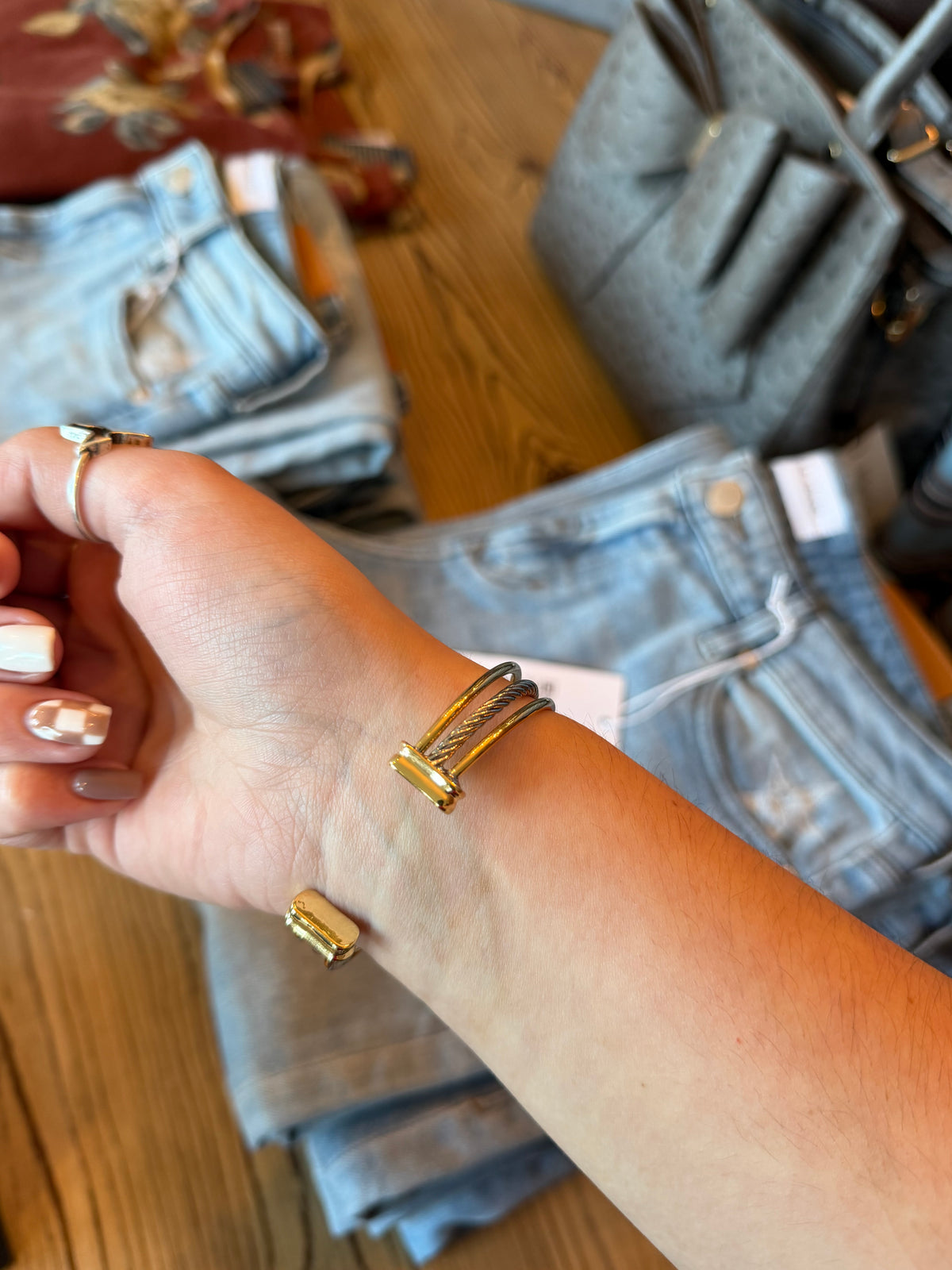 A close-up of a person's wrist wearing a gold-toned wrap bracelet, with folded blue jeans and a gray handbag in the background on a wooden surface.