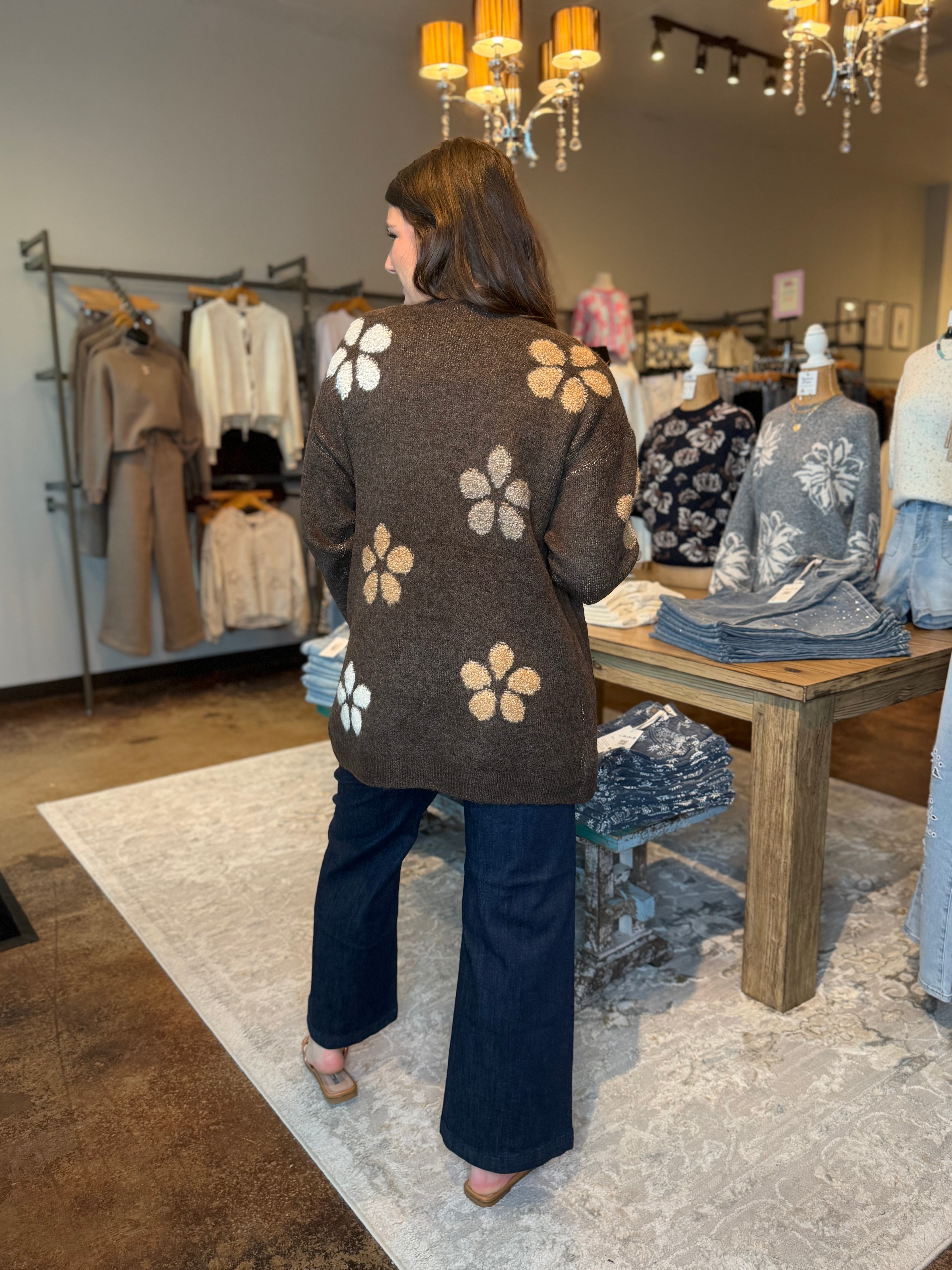 A woman viewed from behind wearing a brown knit cardigan with large white and tan floral motifs, paired with dark wide-leg pants in a boutique clothing store.