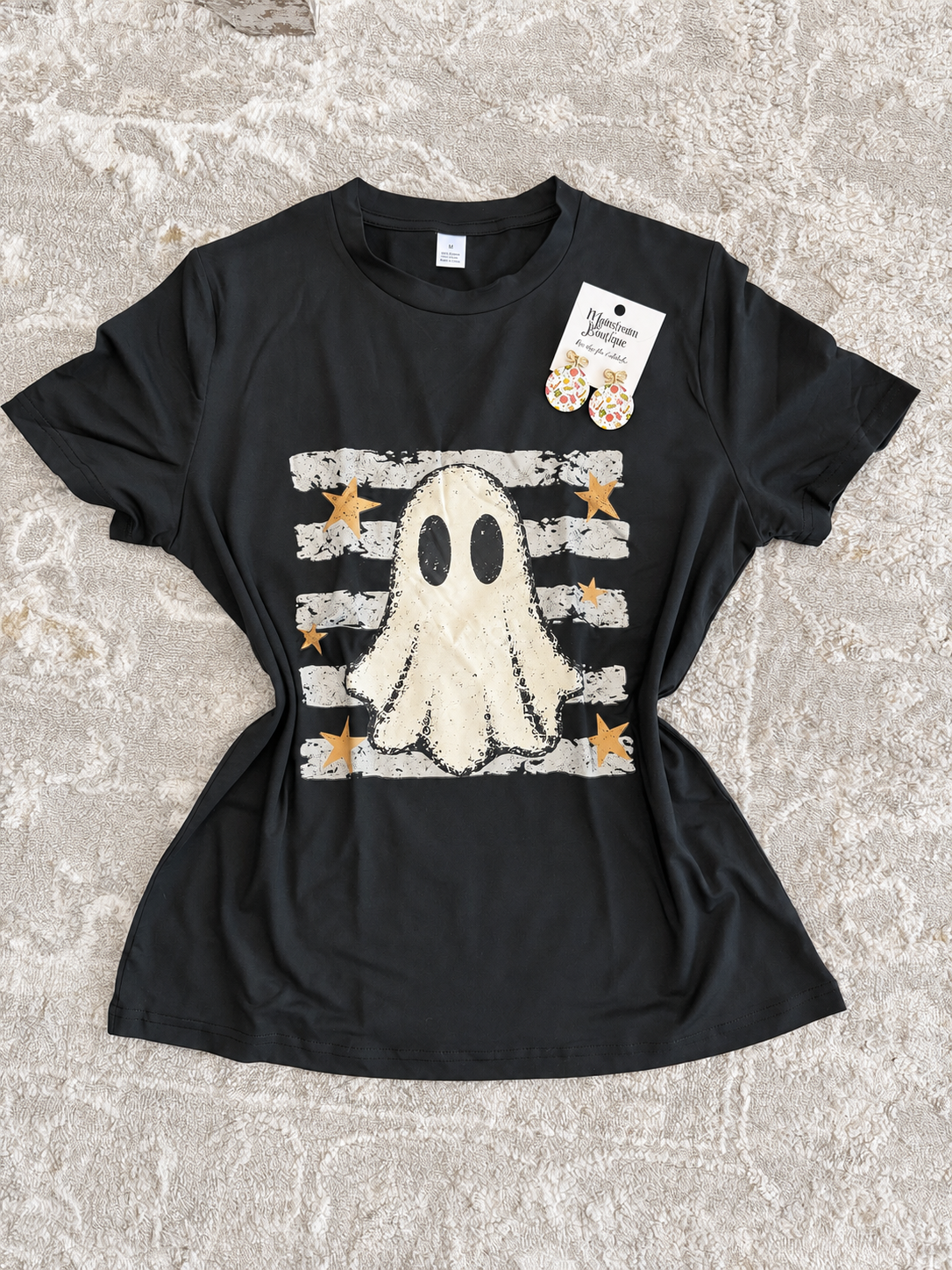 A black short-sleeve graphic T-shirt laid flat on a textured beige rug. The shirt features a distressed white ghost graphic over horizontal white stripes with small gold stars.