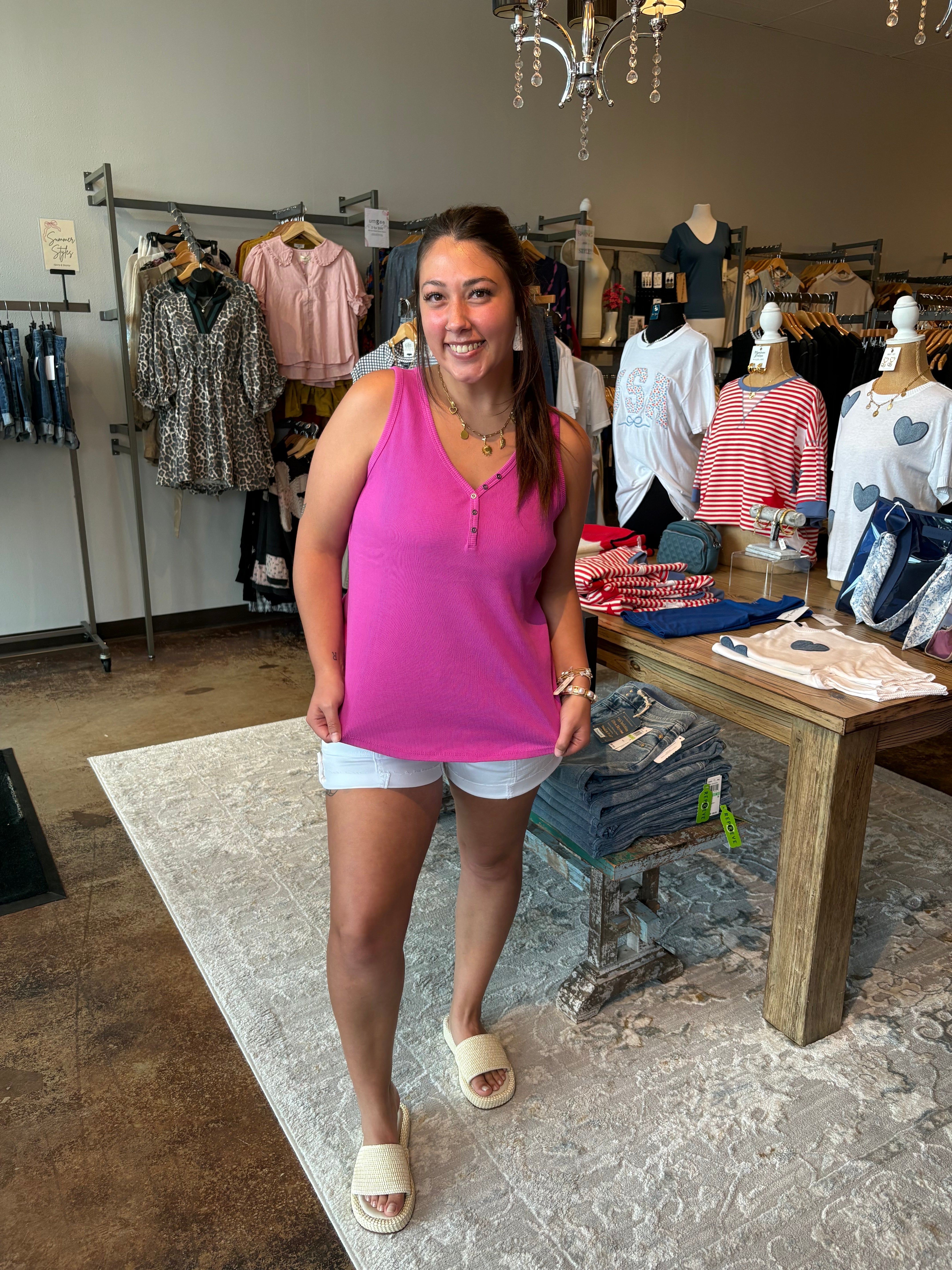 Woman in a pink tank top standing in a clothing store with racks of clothes in the background.