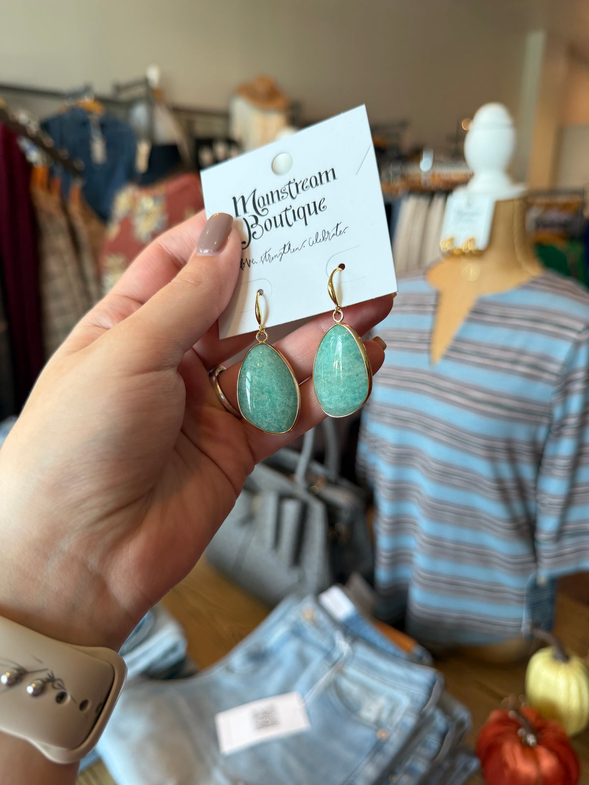 A hand holds a pair of gold hoop earrings with teardrop-shaped light green stone pendants on a white display card labeled 'Monsieur's Boutique'. The background shows a clothing boutique interior with racks of clothes and a mannequin wearing a striped shirt.