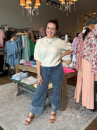 A woman posing in a boutique store wearing a cream-colored textured short-sleeve top and blue wide-leg cuffed jeans.