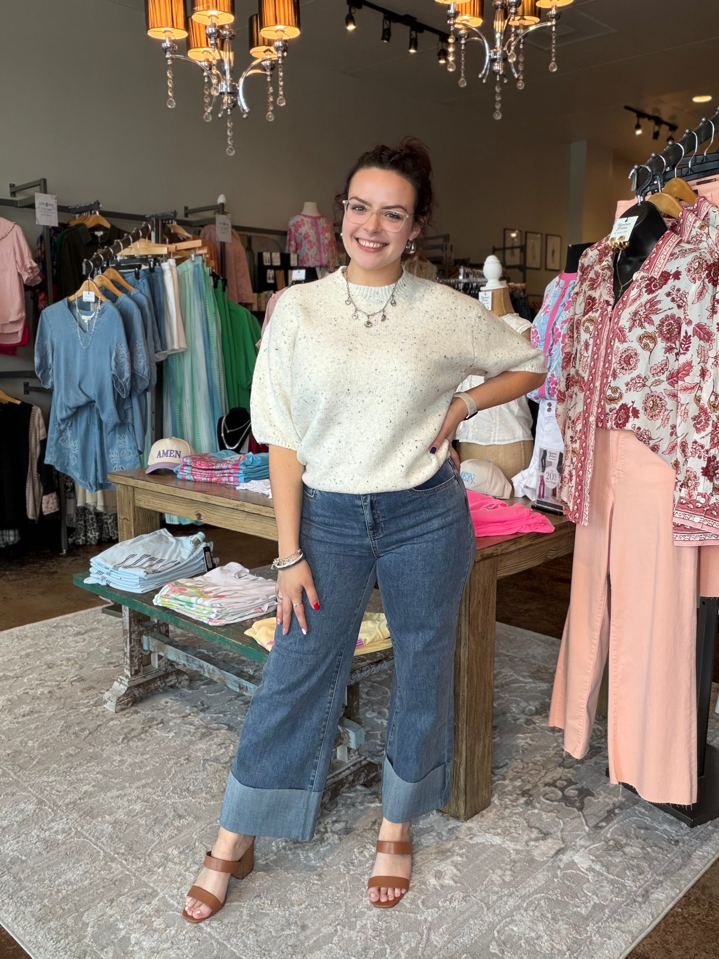 A woman posing in a boutique store wearing a cream-colored textured short-sleeve top and blue wide-leg cuffed jeans.