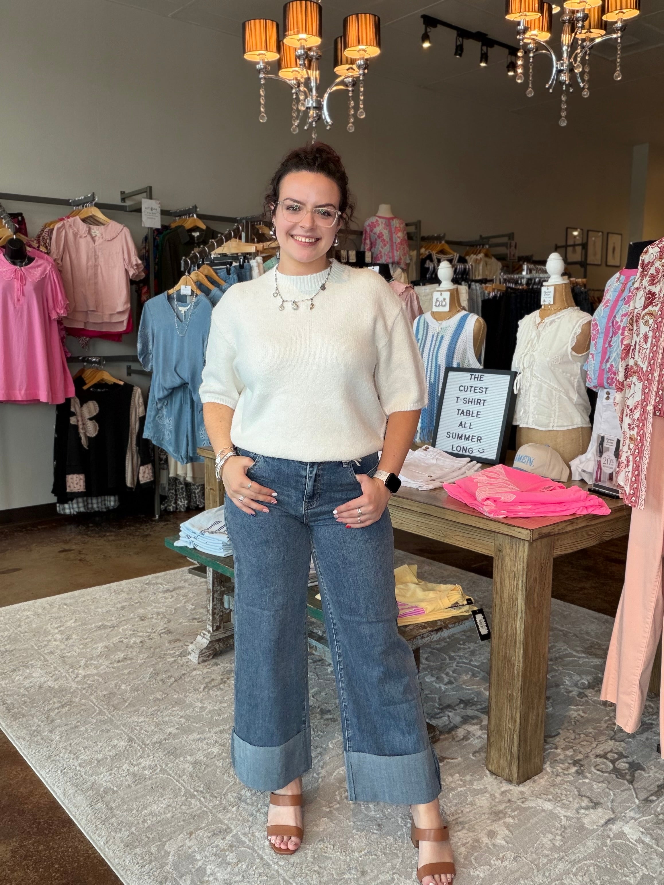 A woman stands inside a clothing boutique wearing a cream short-sleeve knit top and wide-leg blue jeans with rolled hems. The boutique has clothing racks, mannequins, a wooden display table, and hanging chandeliers in the background.