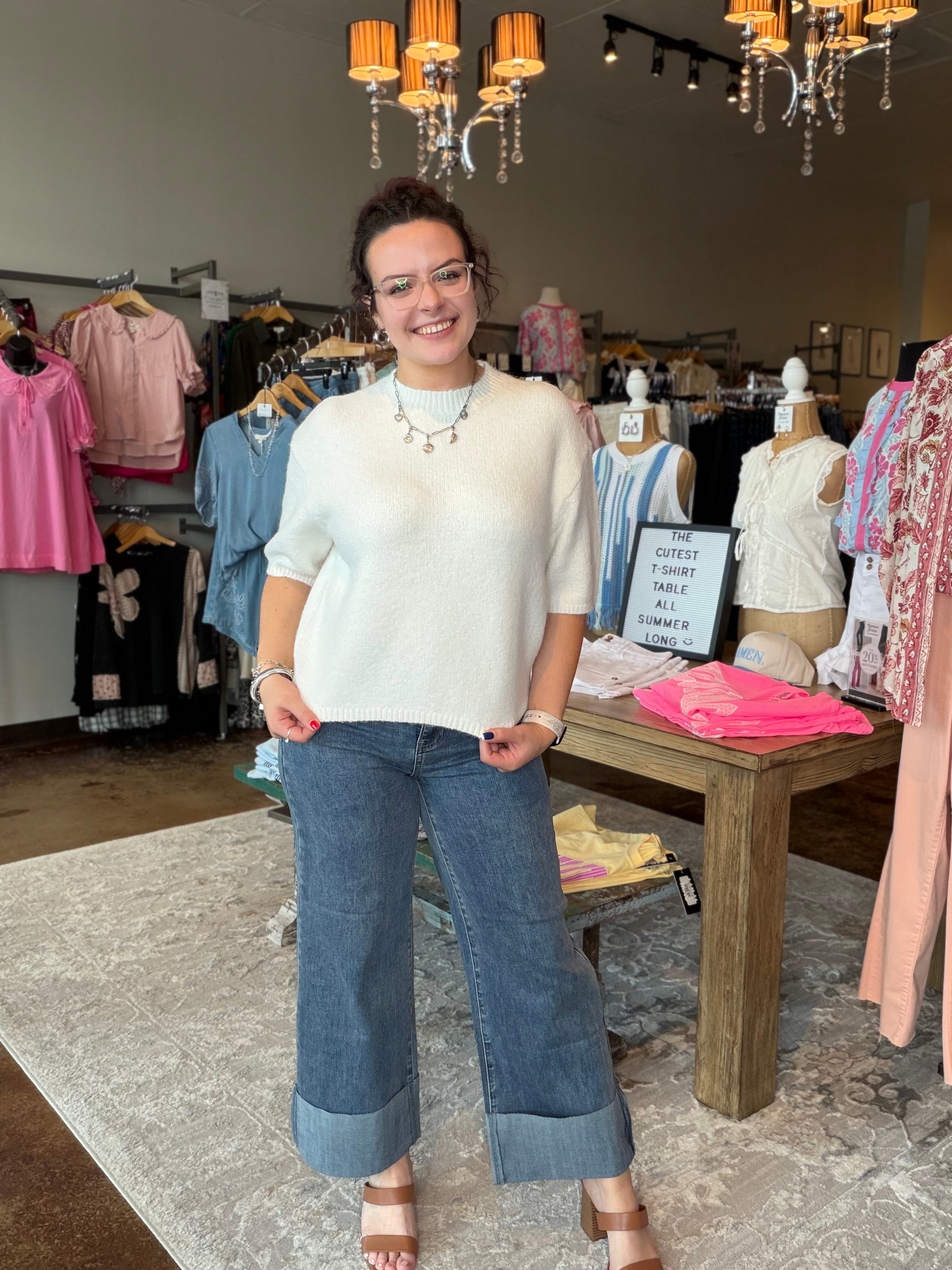 A woman stands in a clothing boutique wearing a cream knit short-sleeve sweater and wide-leg cuffed jeans. The boutique interior features clothing racks, mannequins, a wooden table with folded garments, and hanging chandeliers.