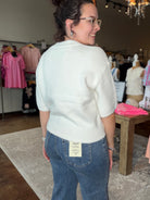 A woman wearing a cream-colored short-sleeve knit sweater and blue jeans, photographed from behind inside a clothing boutique. The back of the jeans features a visible cream brand label that reads 'Cactus'.