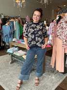 A woman modeling a black short-sleeve floral print top and blue wide-leg jeans with cuffed hems, standing inside a clothing boutique.