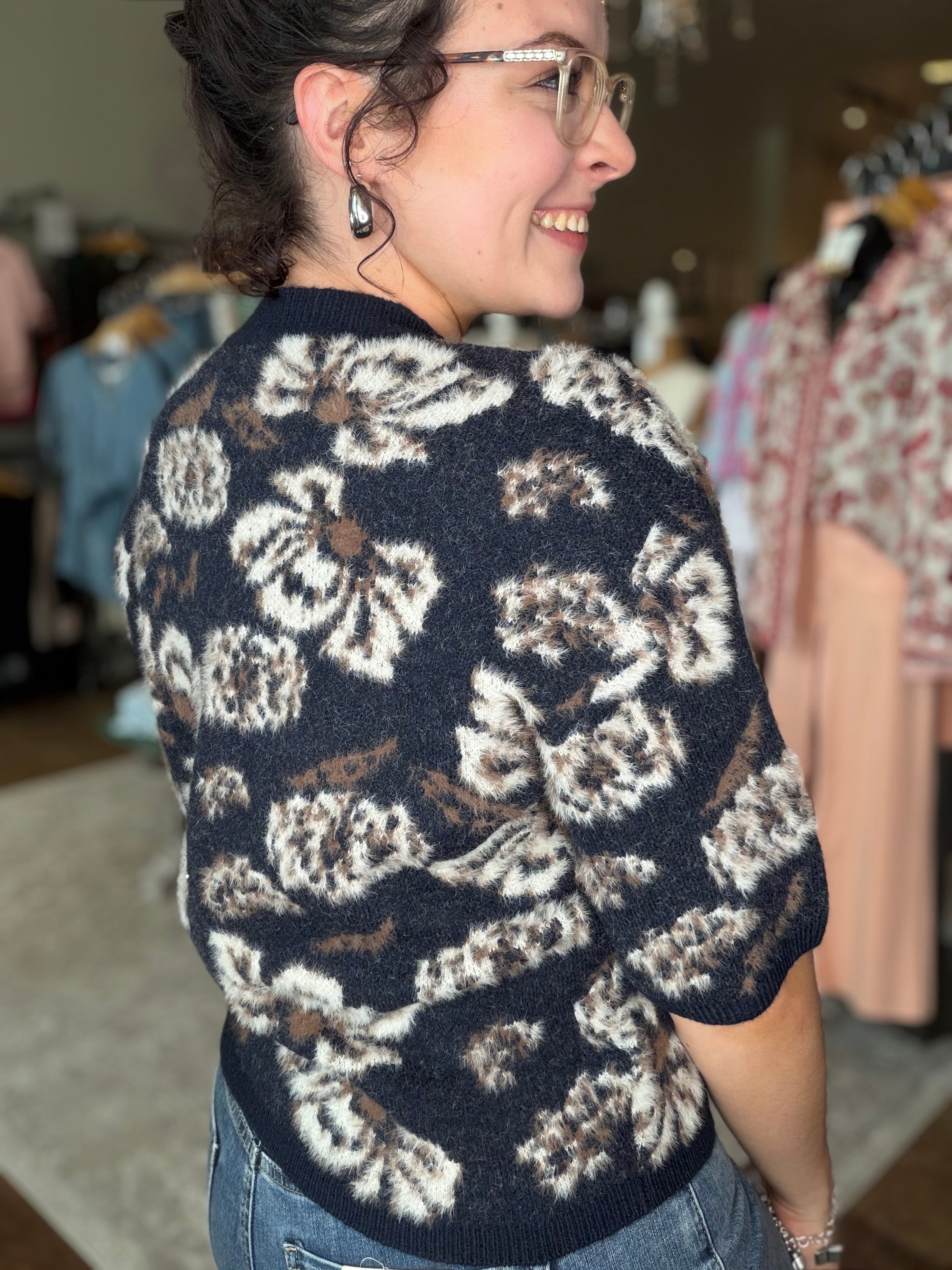 A woman wearing a short-sleeve knit sweater with a large floral pattern in cream, brown, and black, photographed in a clothing store.