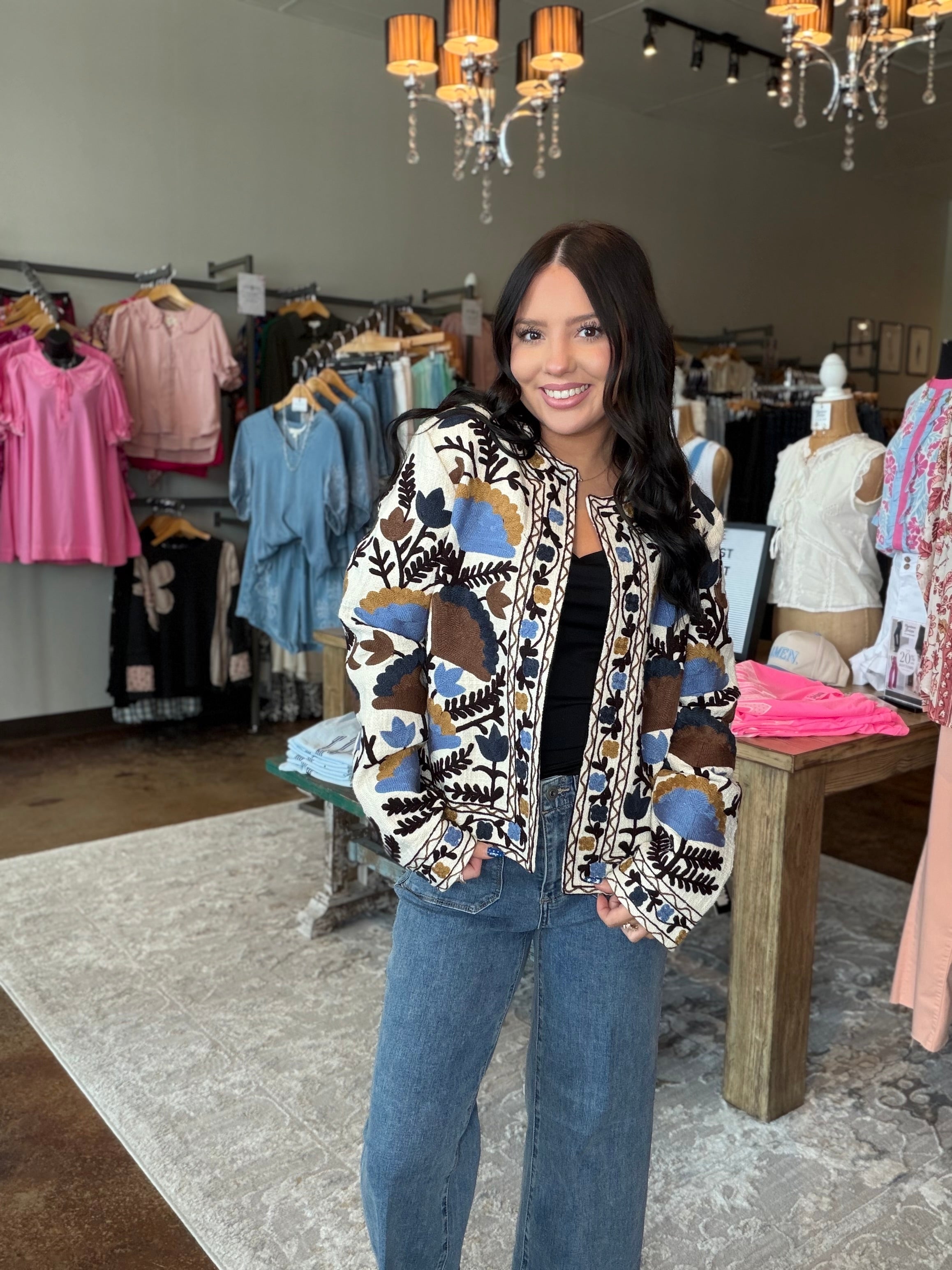 A woman stands inside a clothing boutique wearing a patterned bomber jacket with blue, brown, and beige abstract shapes over a black top and blue jeans.