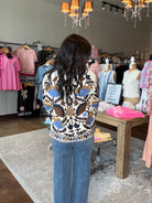 A woman viewed from behind wearing a cream knit sweater with a brown, blue, and black geometric pattern, paired with blue jeans, standing inside a clothing boutique.