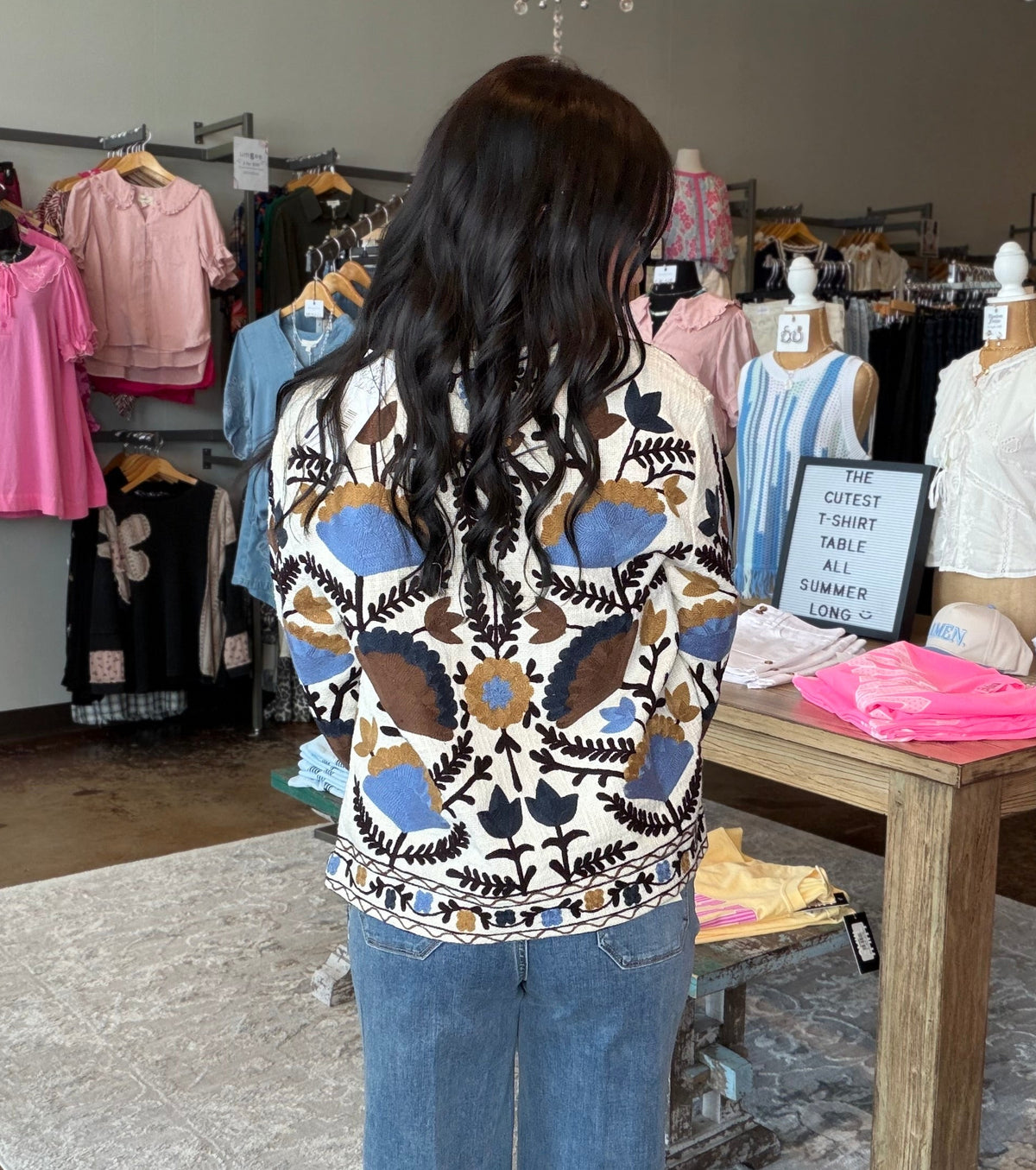 A woman viewed from behind wearing a cream knit sweater with a brown, blue, and black geometric pattern, paired with blue jeans, standing inside a clothing boutique.