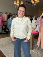 A woman wearing a cream cable-knit long-sleeve sweater and blue jeans stands in a clothing boutique.