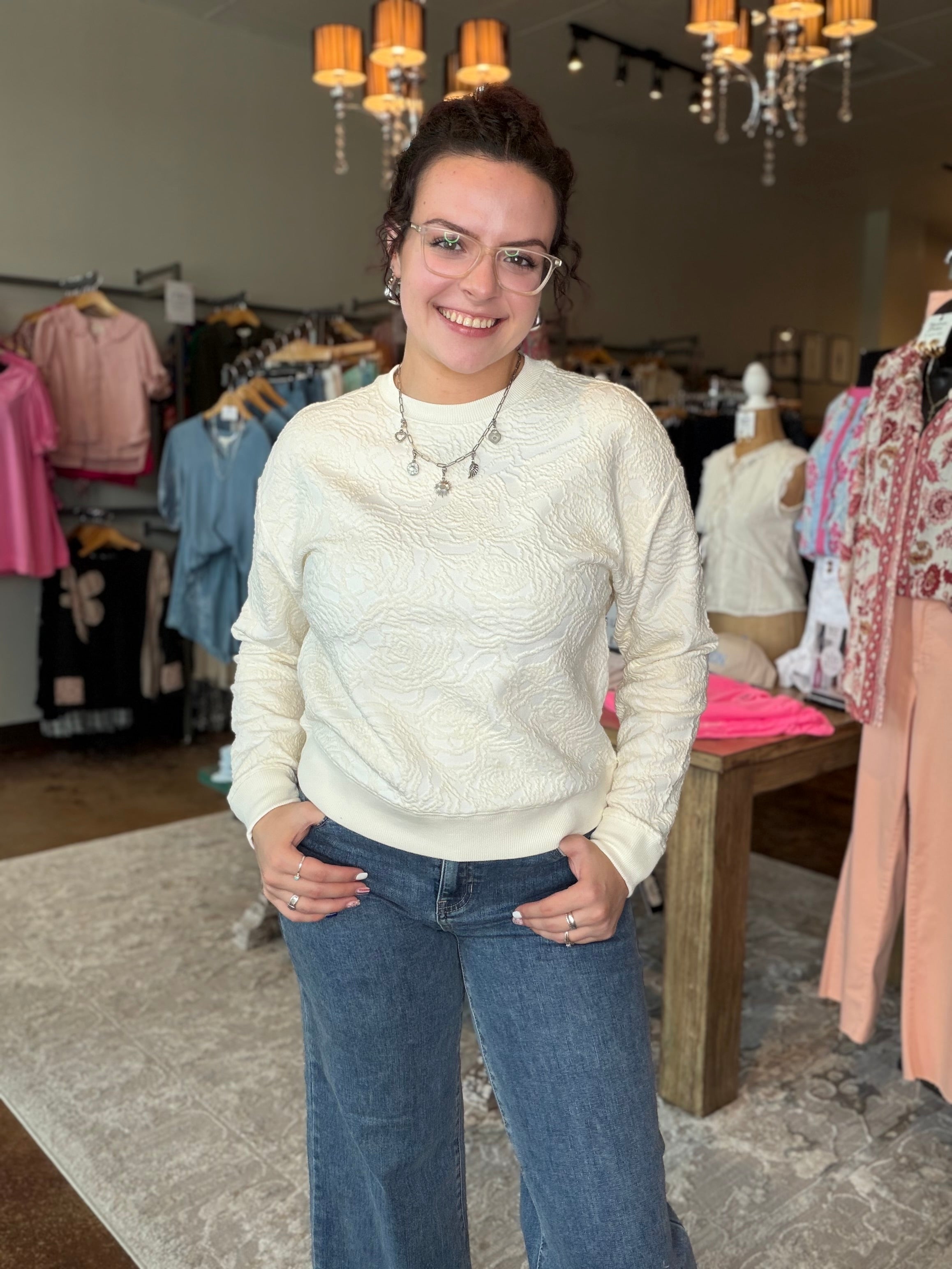 A woman wearing a cream cable-knit long-sleeve sweater and blue jeans stands in a clothing boutique.