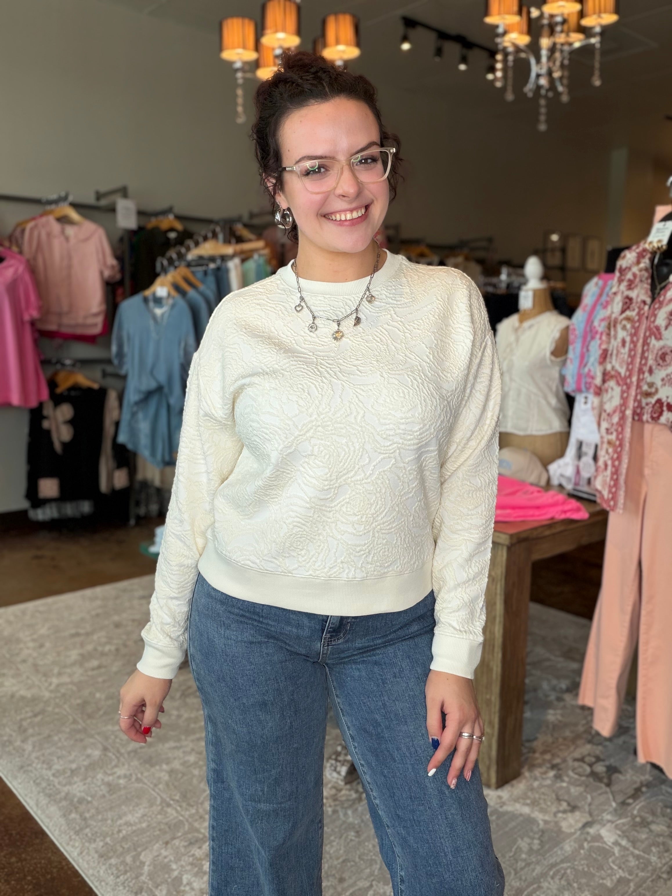 A woman wearing a cream-colored textured knit sweater and blue jeans stands inside a clothing boutique.