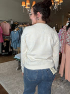 A woman wearing a cream-colored textured knit cardigan and blue jeans, photographed from behind inside a clothing boutique.