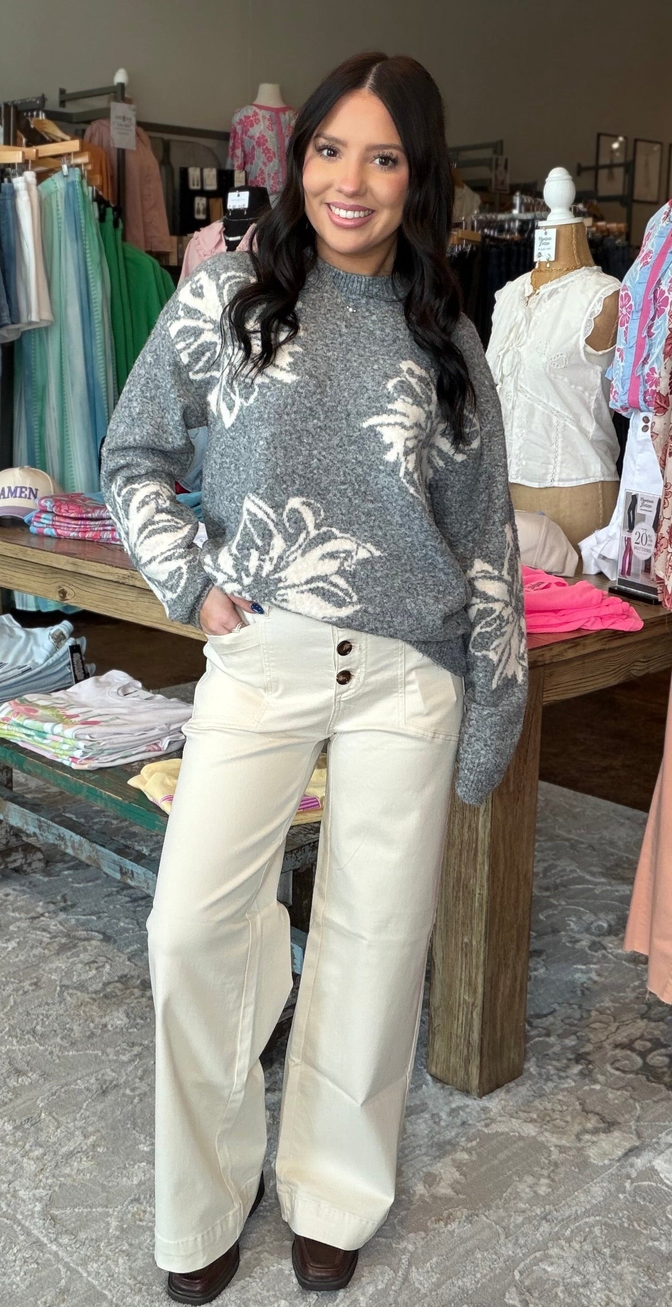 A woman stands in a clothing boutique wearing a gray floral knit sweater and cream wide-leg pants. Clothing racks, mannequins, a chandelier, and a patterned rug are visible in the store interior.